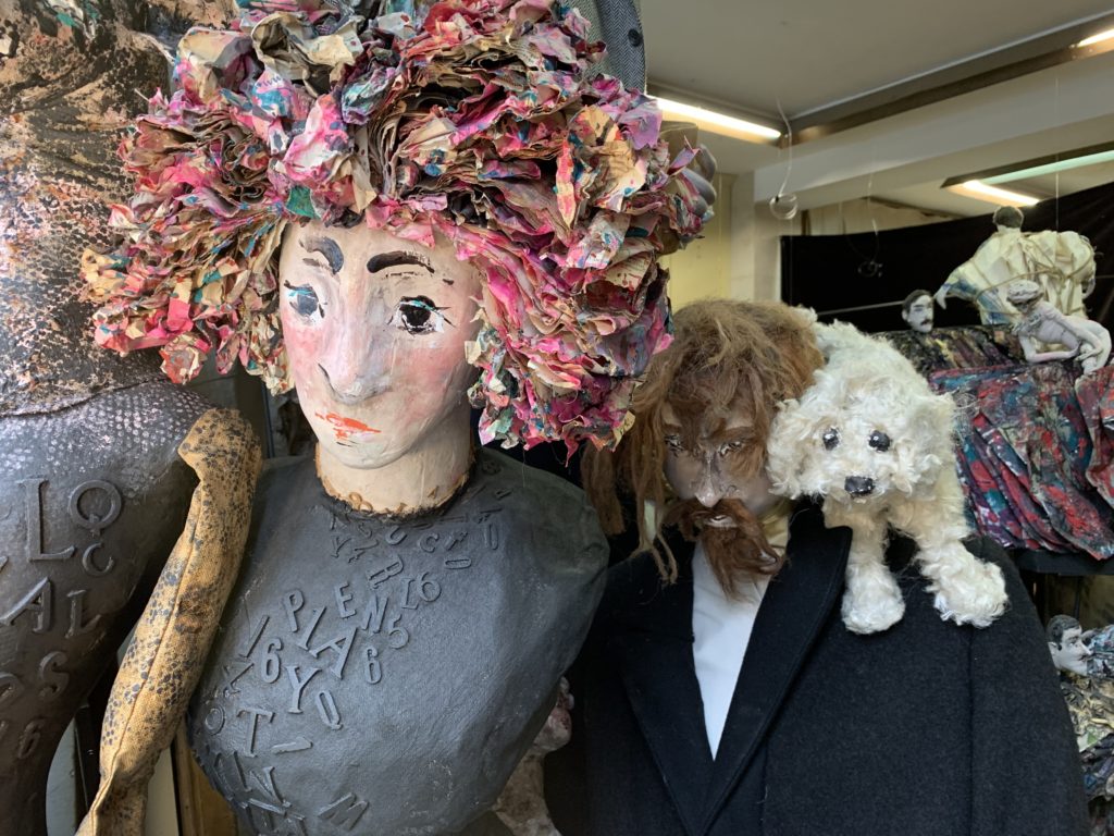 tête de femme en papier maché avec une perruque de papiers rose, homme à tête de chien avec un petit chien blanc sur l'épaule