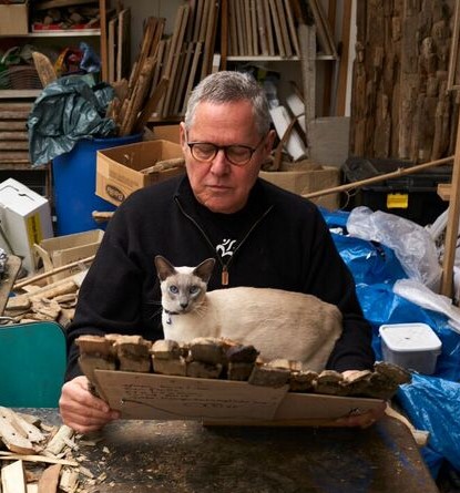 photographie de Marc Bourlier dans son atelier entouré de bois flottés et oeuvres en compagnie de son chat siamois, recadré