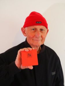 photographie couleur de l'artiste Pierre Albasser portant un bonnet rouge et montrant un de ses livres d'artiste façon leporello