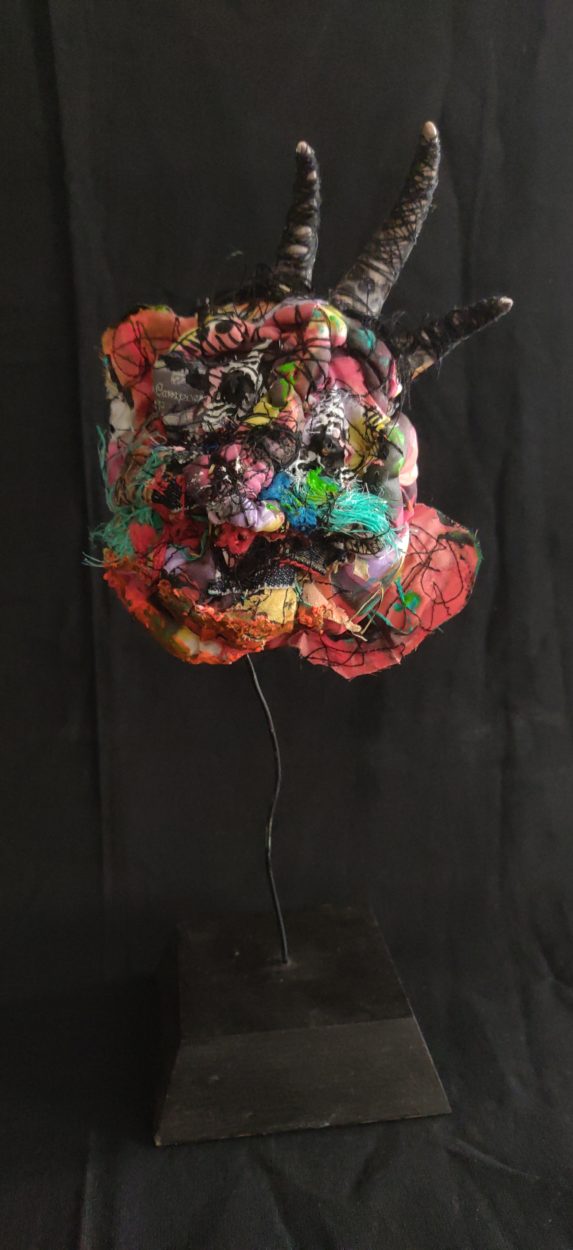 Image qui représente une tête en sculpture textile peinte aux diverses techniques , sur une tige en métal et un socle en bois peint en noir