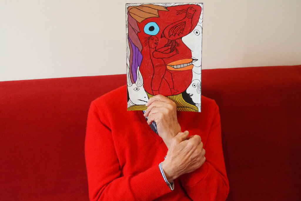 photo qui représente l'artiste Pierre Albasser en pull-over rouge assis sur un canapé rouge qui tient une oeuvre devant son visage